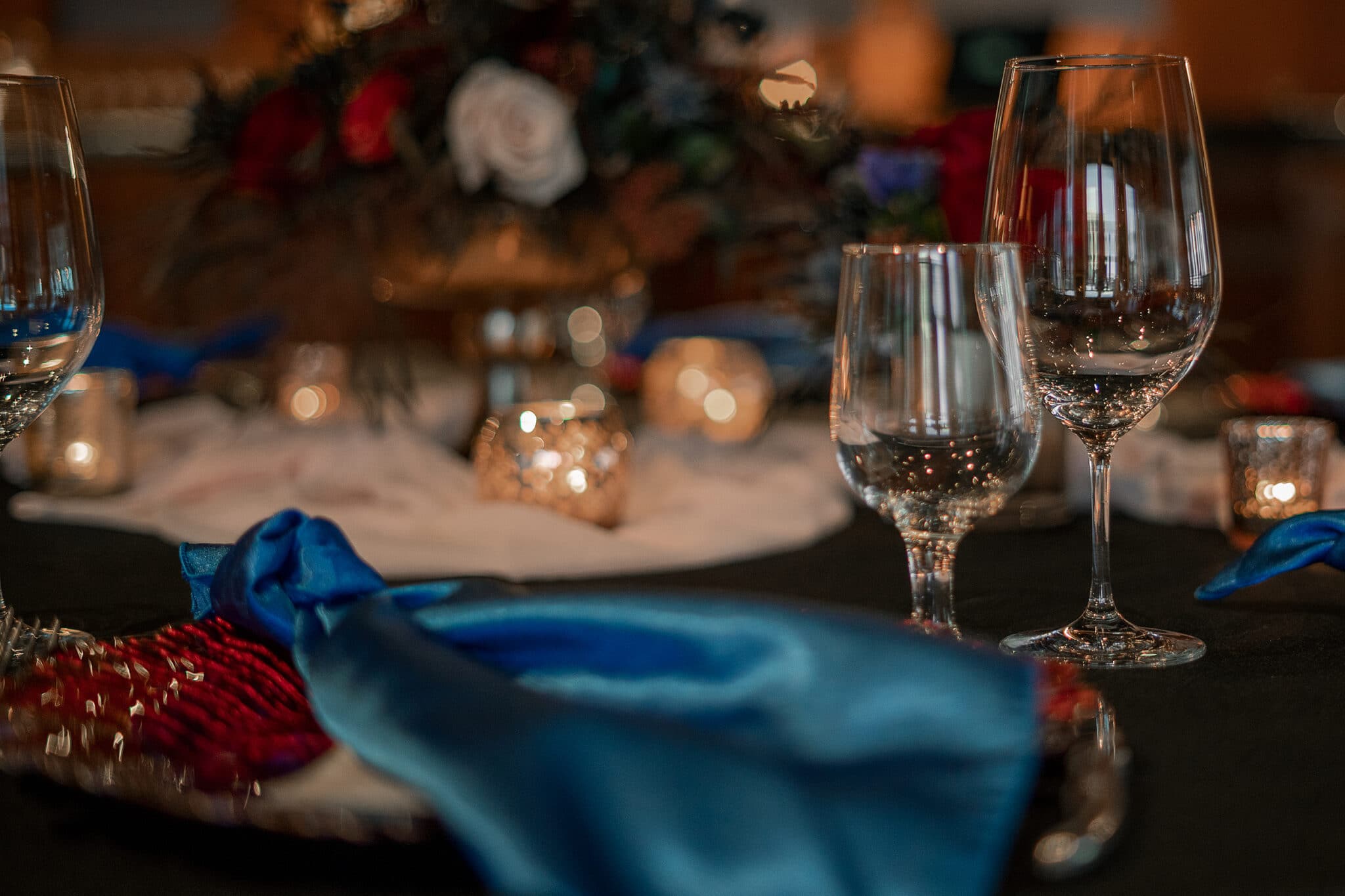 A table set awaiting the start of an event, decorated with red charger plates, blue napkins, candles, and a bouquet of roses, pictured close up