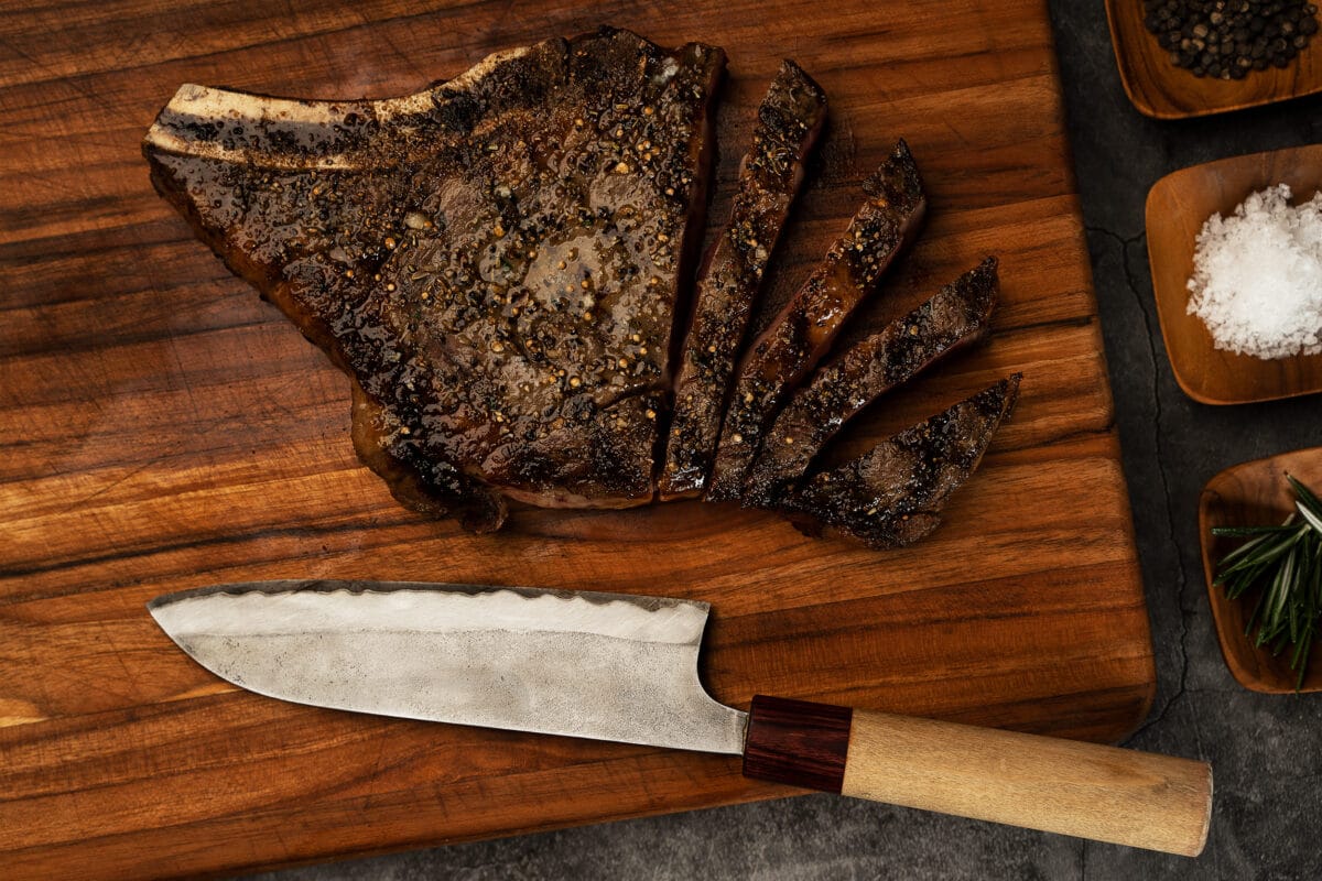 An 18ox. ribeye steak on a cutting board, partially sliced, with a steak knife pictured beside it.