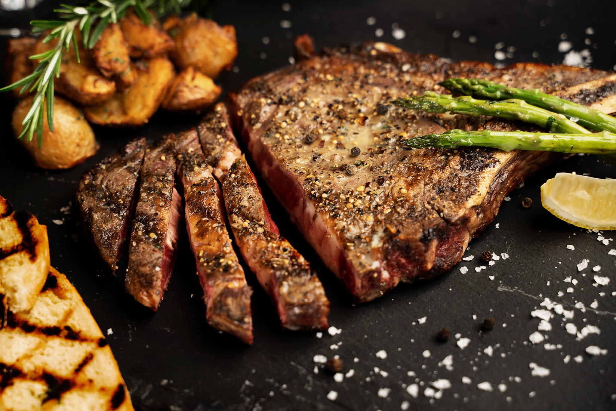 A closeup of a ribeye steak, well seasoned and medium rare, partially cut into strips, with sides of asparagus, potatoes and char-grilled toast.