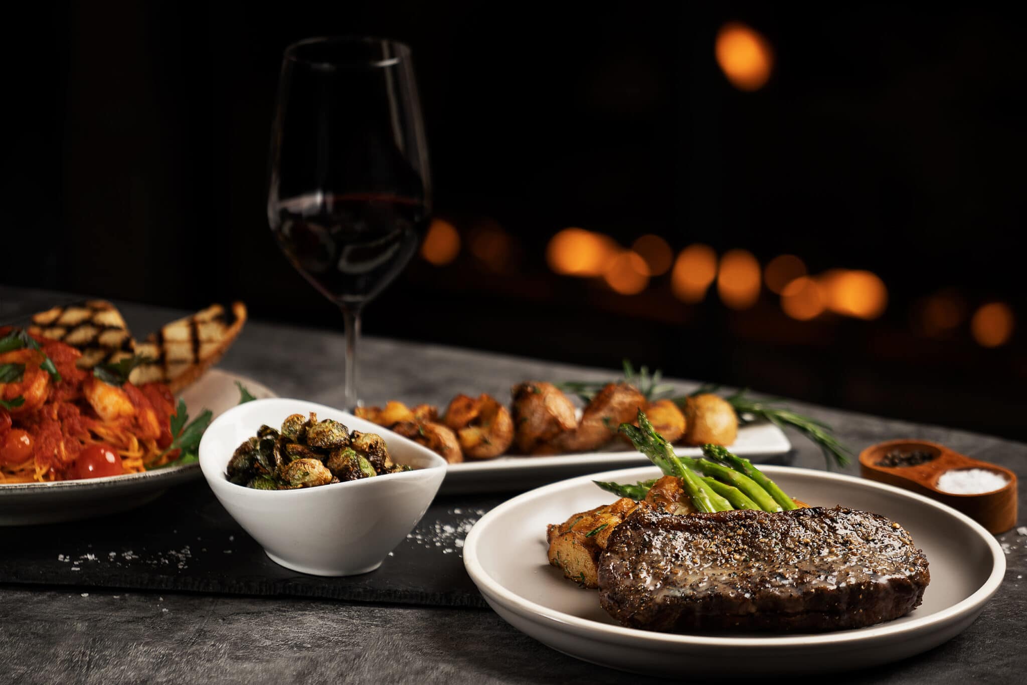 A dark and moody photo of the Satlik menu, steak in the foreground, sides and a glass of red wine in the background