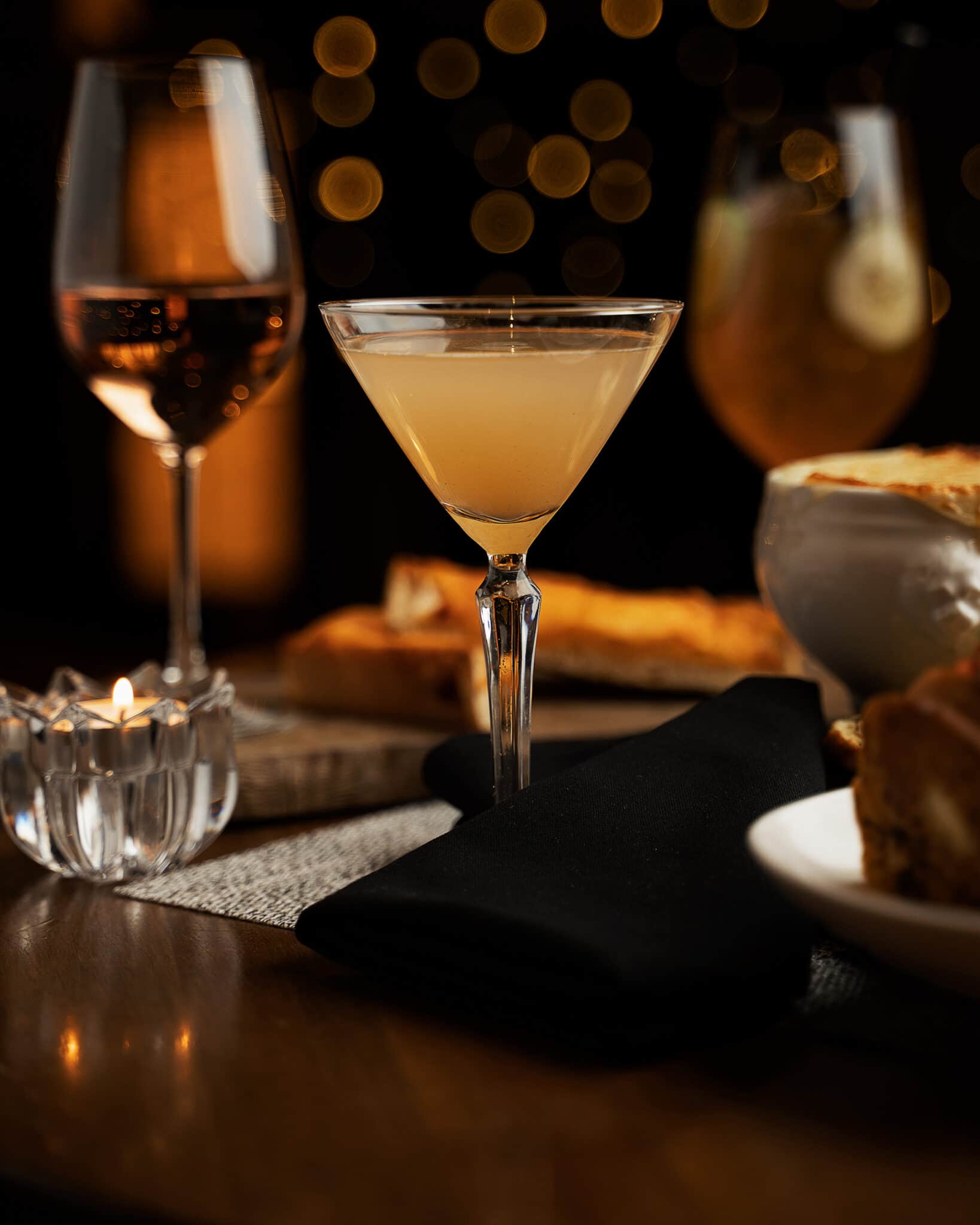 A dark and moody photo of a martini, with a glass of wine and a french onion soup in the background