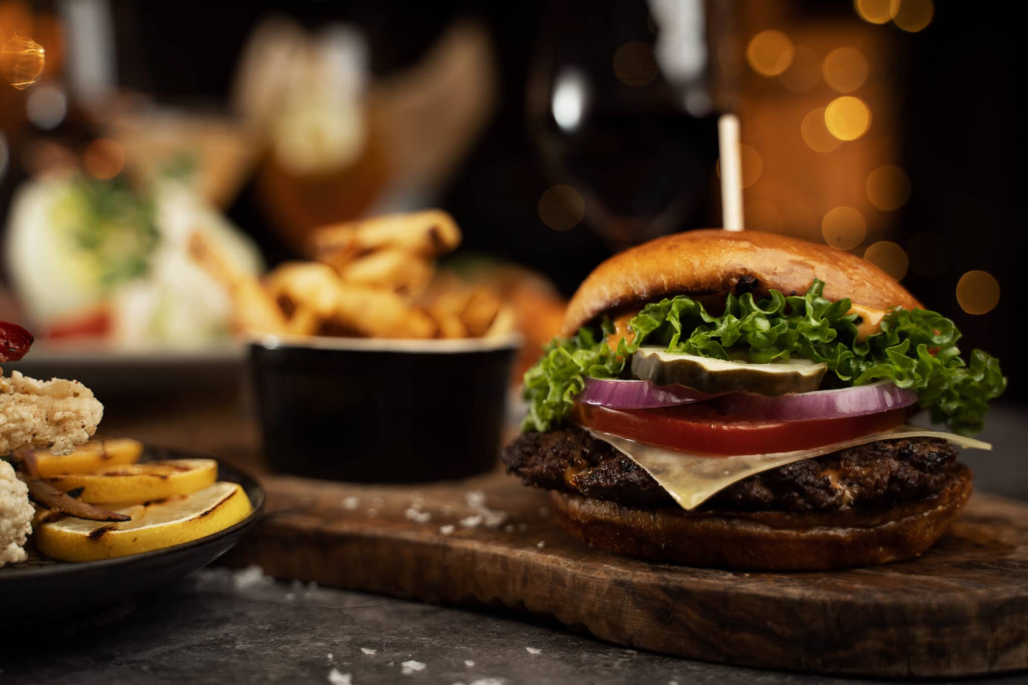 A dark and moody photo of burger on a wood serving board, with sides
