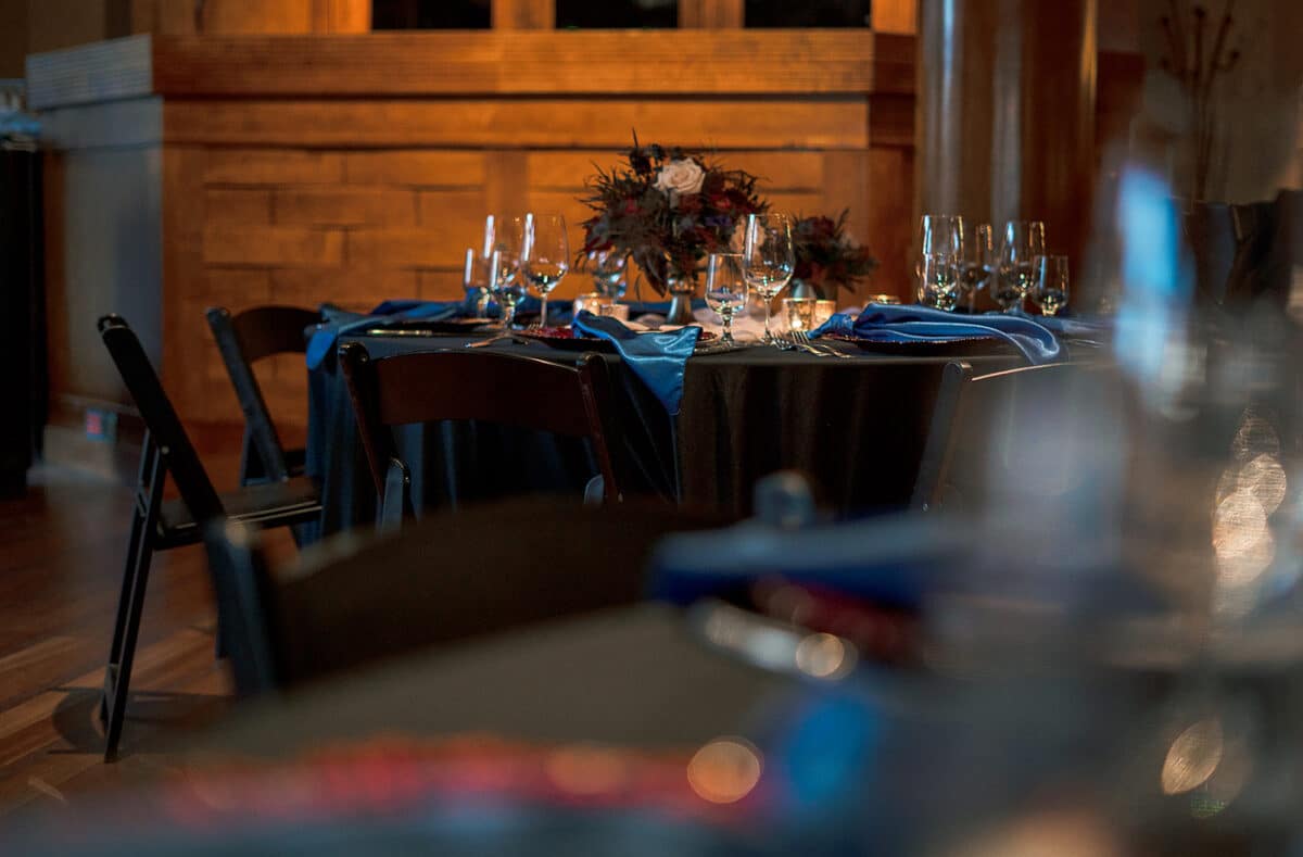 A table set awaiting the start of an event, decorated with red charger plates, blue napkins, candles, and a bouquet of roses, pictured from a distance