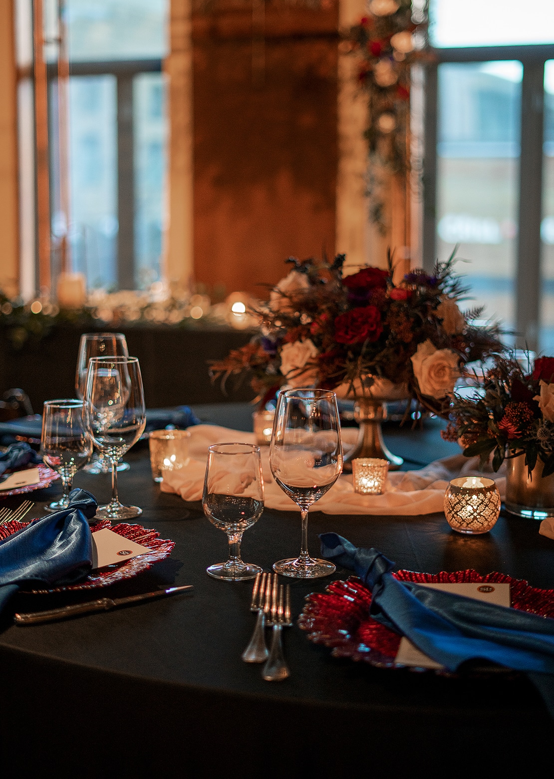 A table set awaiting the start of an event, decorated with red charger plates, blue napkins, candles, and a bouquet of roses