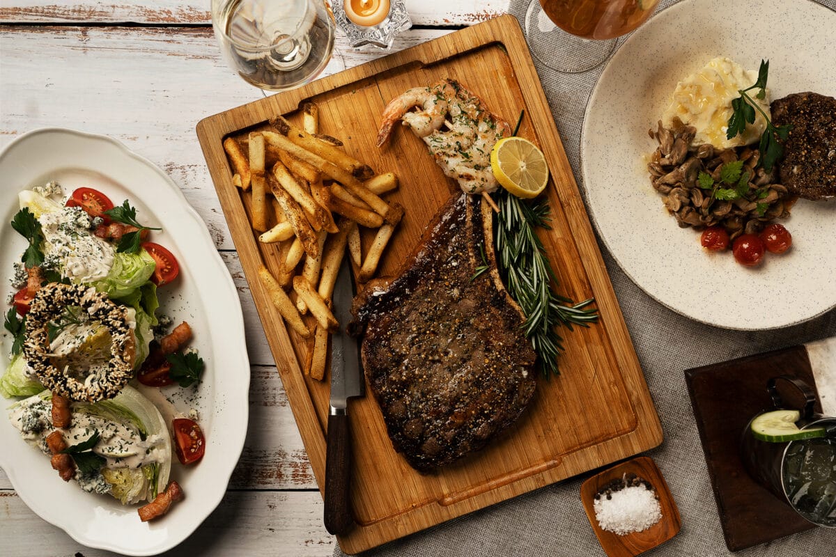 Saltlik 18oz ribeye on a cutting board with shrimps and french fries, with a prime sirloin entree to the right and the side-salad to the left.