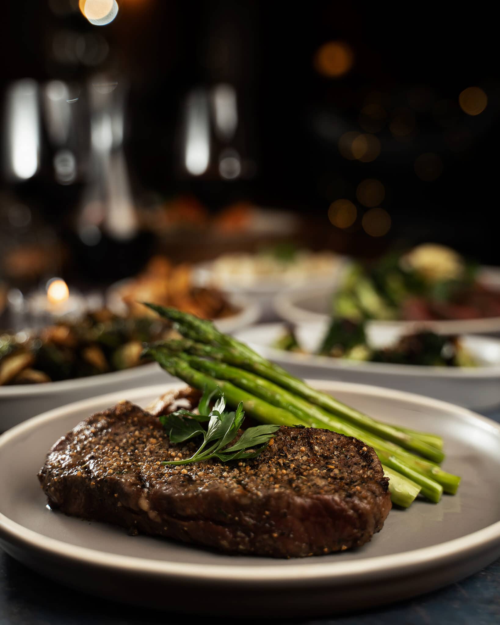 A steak with asparagus on a table with other dishes and glasses of wine in the background